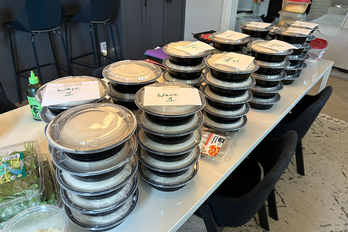 Photo of the 11 stacks of individual pizza dough rounds (5 per stack) that Joe prepped ahead of the Pizza Night event. Each pizza dough round is sealed in a black plastic, take-out-type salad container and the stacks are marked with pieces of paper labeled "Wave 1," Wave 2," etc.