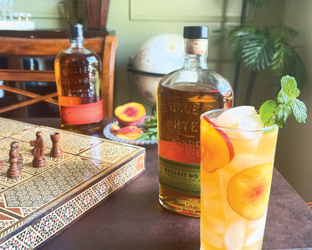 Give Peach A Chance bourbon drink in a tall glass with Bulleit Bourbon bottle in the background.