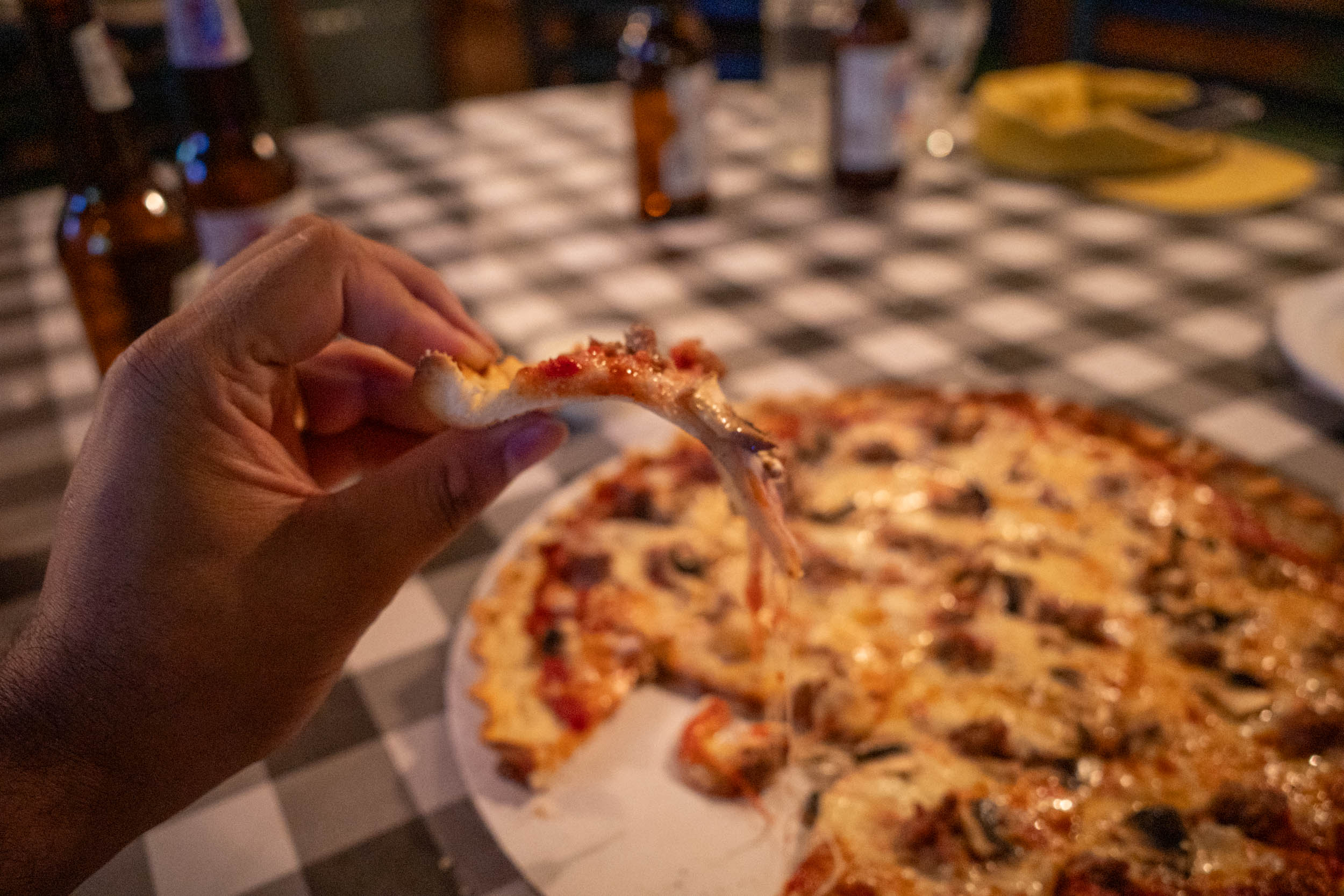 hand holding pizza slice with full pizza in the background on checkered tablecloth