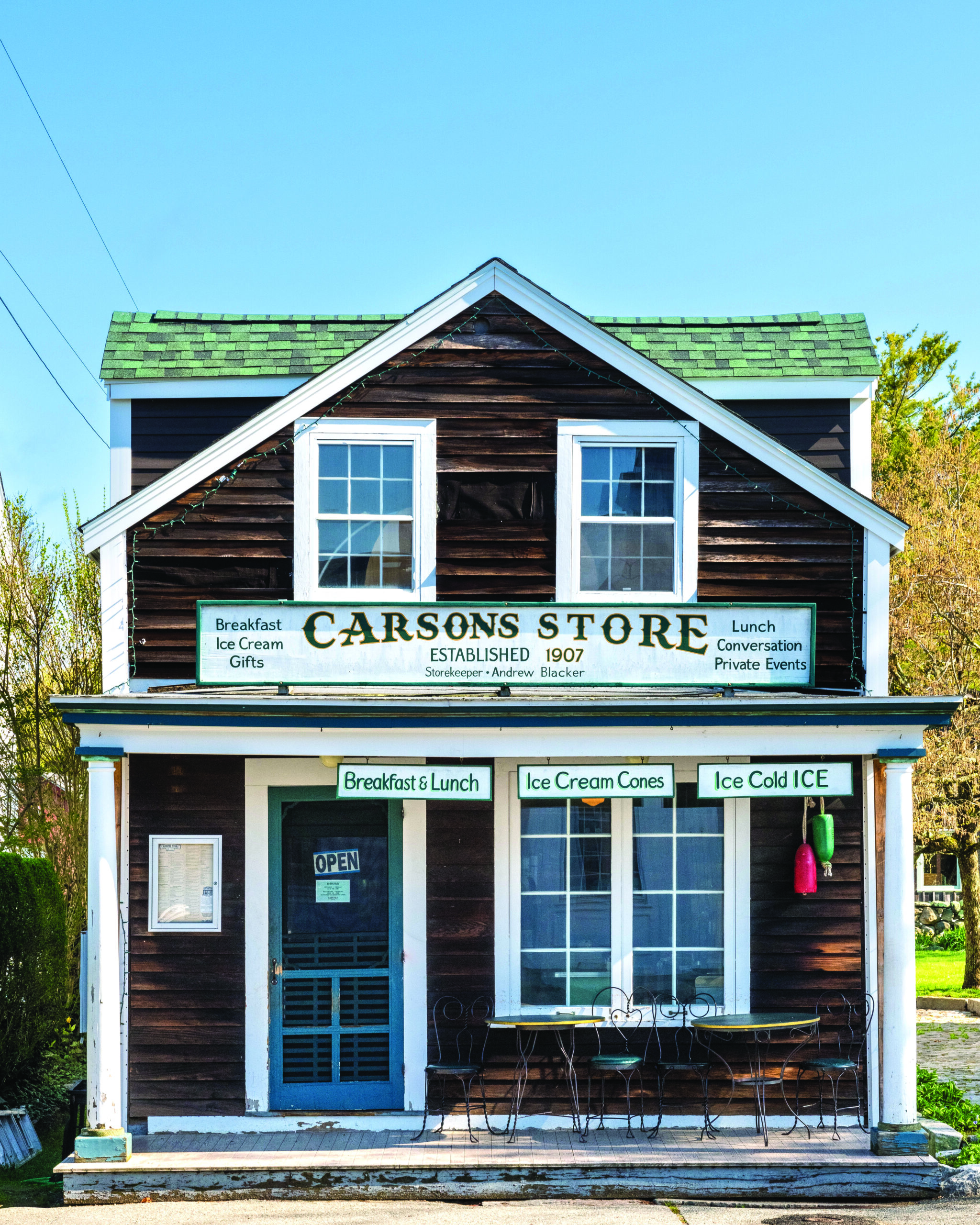 dark wooden sided building with white trim and details. Sign above reading "Carson's store", sells food and ice cream. Small metal tables and chairs on the porch.  