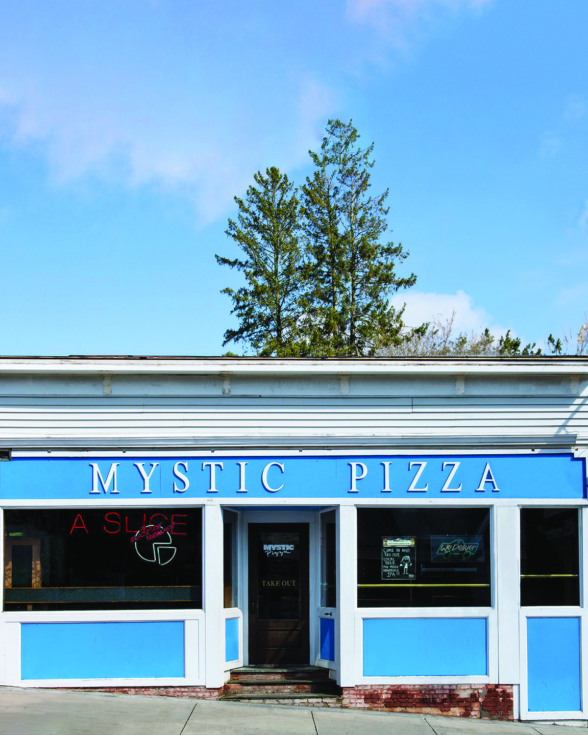 photograph of mystic pizza restaurant storefront. There is a blue sign above the door and neon signs in the window. the sky in the background is clear and blue. 