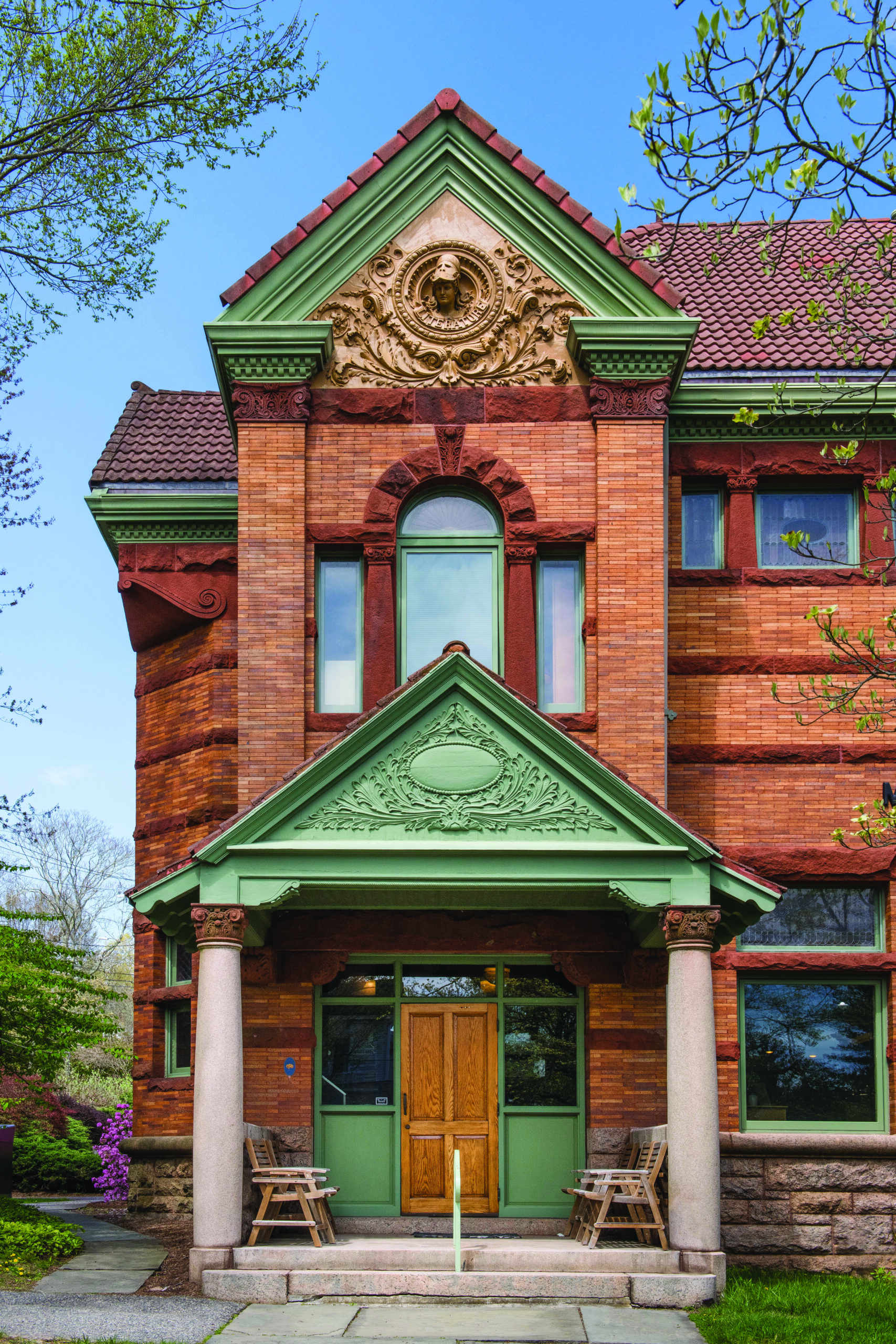 old ornate building entry way. red brick with sage green trim and detailing. there is a wooden door in the front with chairs outside.
