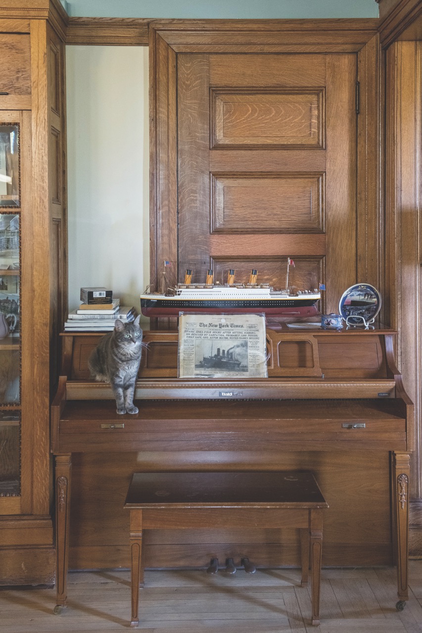 old wooden piano with a model ship and a cat on top of it. wooden trim and door in the bakcground. 