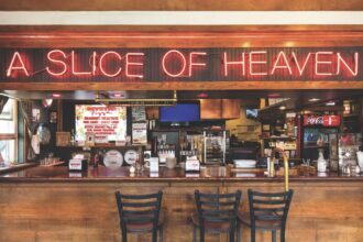 shot of wooden bar at a pizza restaurant with neon red sign above reading "a slice of heaven". There are three darker bar stools in the foreground and a variety of colorful trinkets in the background