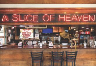 shot of wooden bar at a pizza restaurant with neon red sign above reading "a slice of heaven". There are three darker bar stools in the foreground and a variety of colorful trinkets in the background