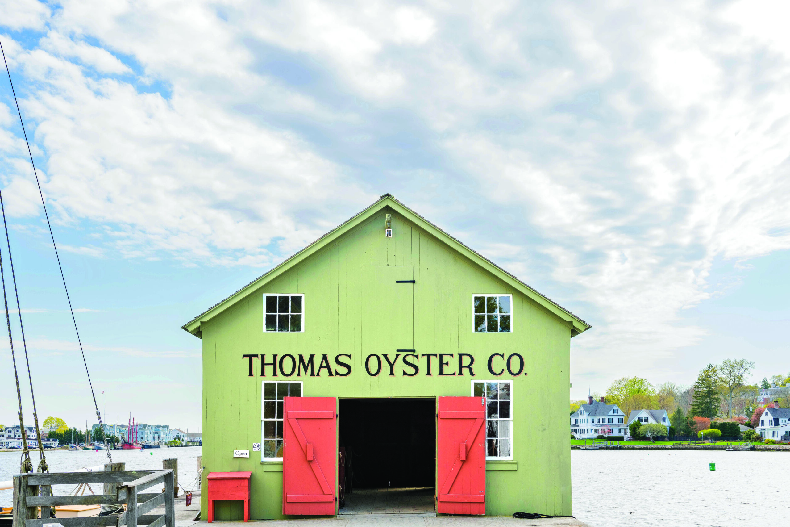 pale green building with red doors and a sign above entryway reading "thomas oyster co." The building has small windows and is situated on a dock. There's ocean and cloudy sky in the background. 