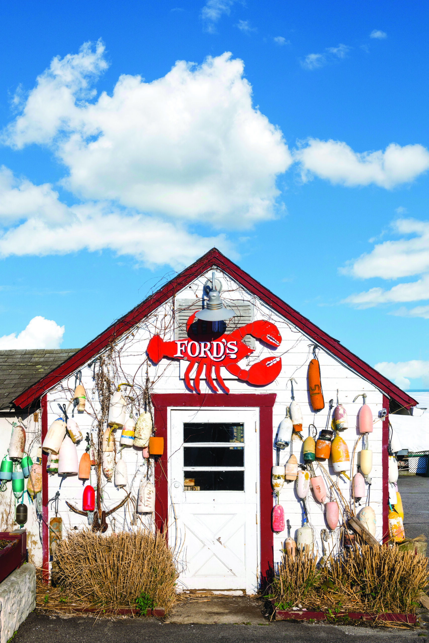 photograph of nautical shed with a lobster sign hanging over it reading "ford's". bright blue sky with white clouds in the background. Bouys hanging on the outside wall. 