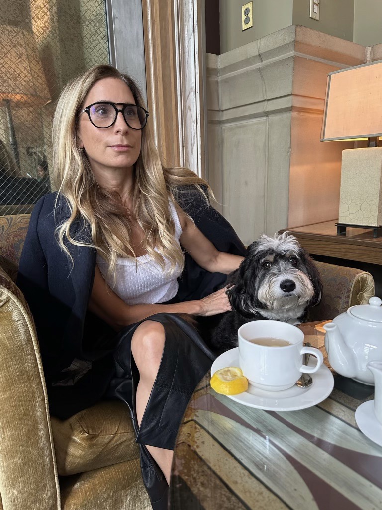 Blonde woman sitting in comfy chair with her dog. She is wearing dark glasses and a blazer. In the foreground there is a coffee table with tea and a lemon slice on it. 