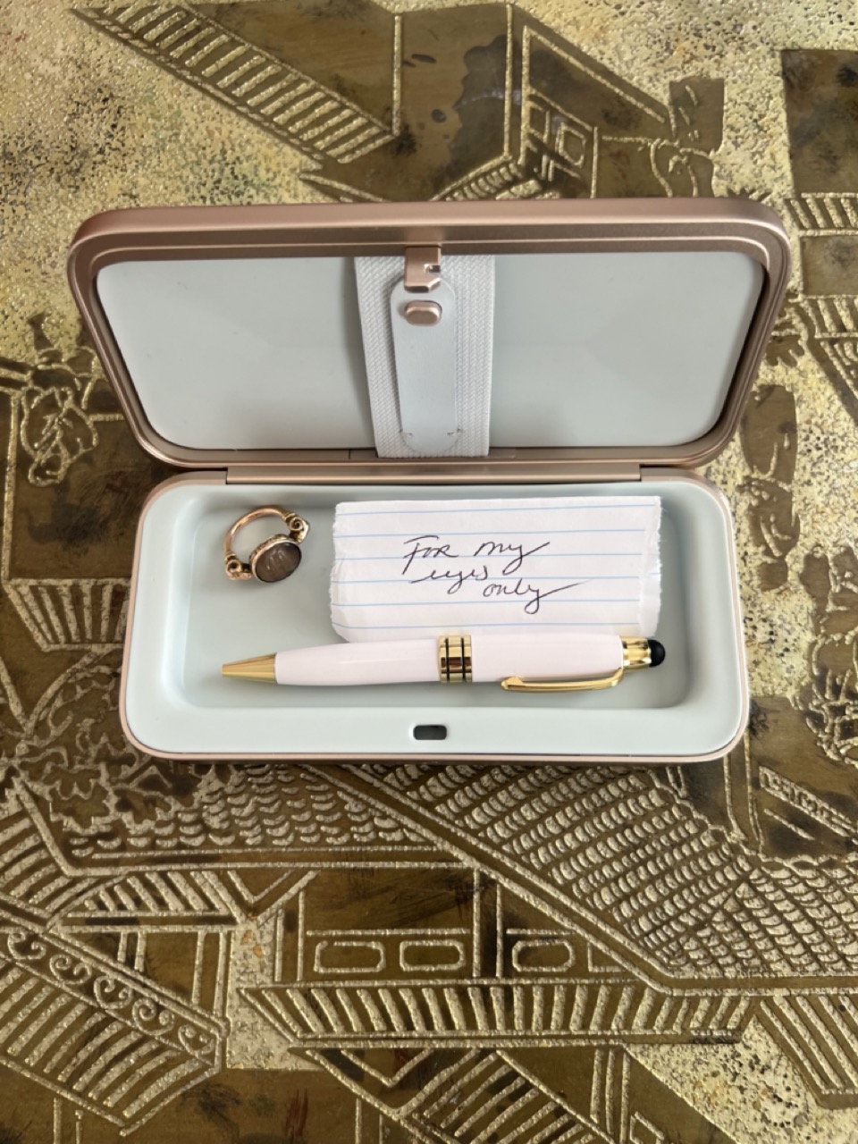 photograph of a TROVA go safe containing a pen, a ring, and a piece of paper on a patterned gold and cream background