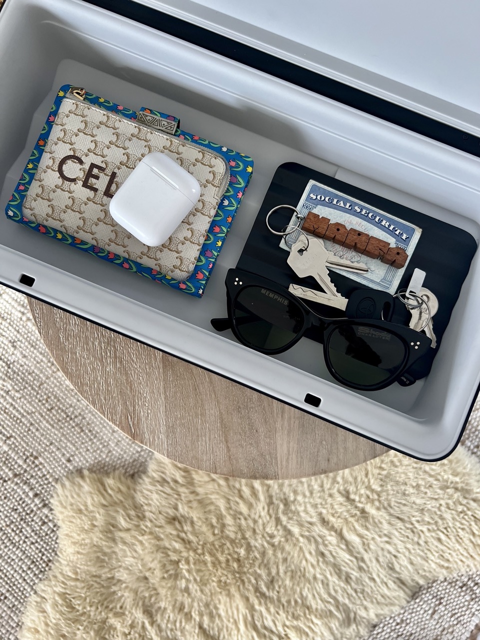close up shot of a TROVA home safe with keys, airpods, sunglasses, a wallet, and more inside. Plain textured carpets in the background 