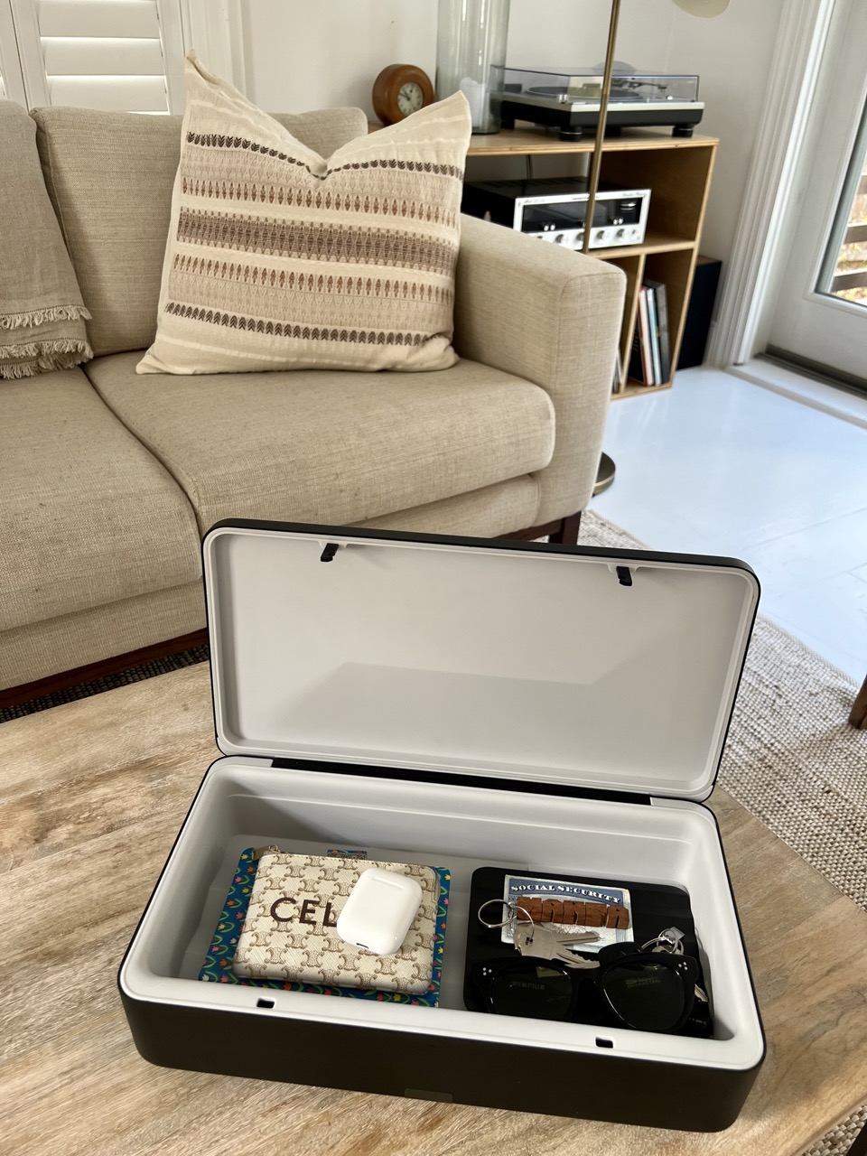 photograph of a TROVA home safe on a coffee table containing a variety of items; keys, AirPods, a journal, a pair of glasses. 