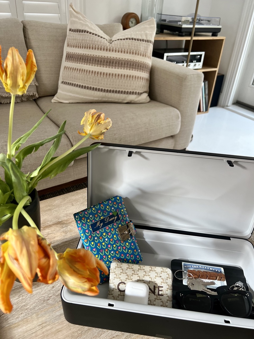 TROVA home safe on a coffee table with keys, airpods, sunglasses, a wallet, and more inside. A jar of fresh flowers are on the table next to it and there is a couch in the background. 
