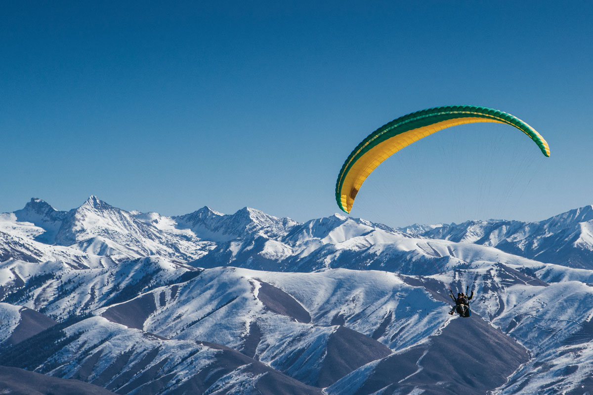 Person paragliding with a green and yellow parachute over snowy covered mountain peaks.