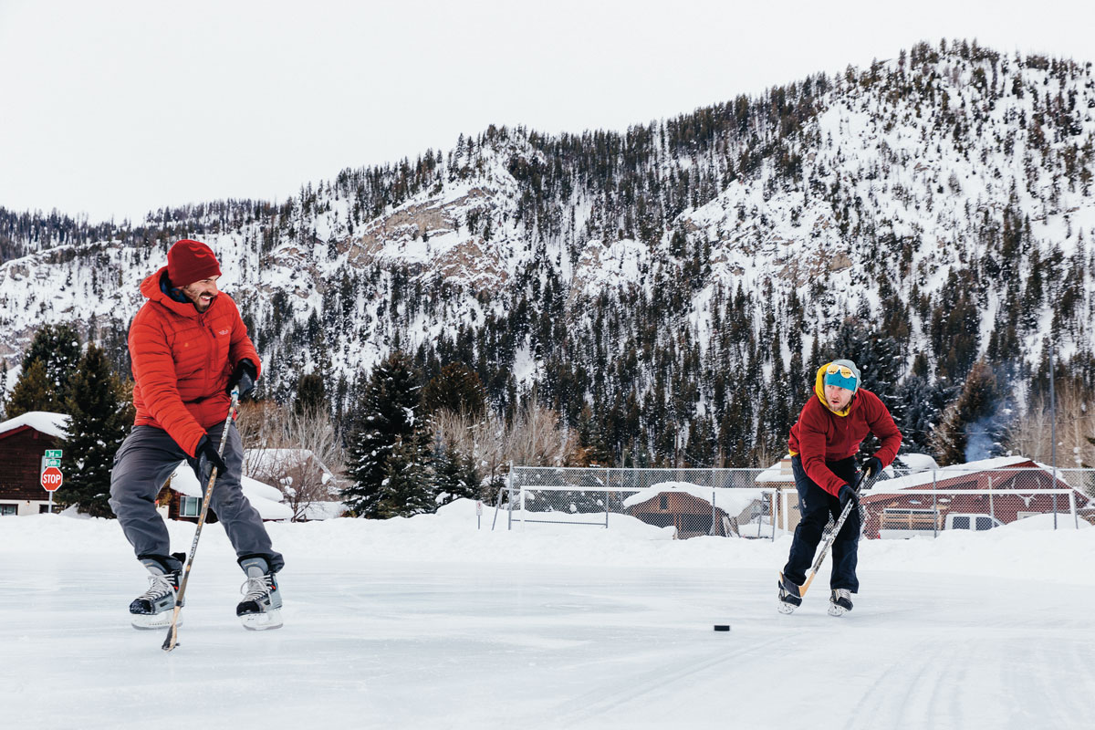 2 people playing hockey outside at the Cristina Potters Outdoor Ice Rink with snowy tree covered mountains in the background