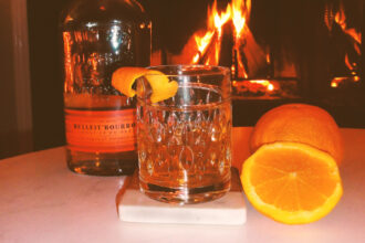 Bottle of Bulleit in front of a fire with a sliced orange and a glass of bourbon.