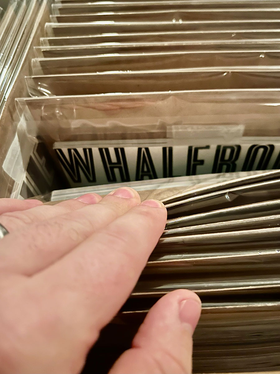 Close-up photo of Matthew Wisotsky's WHalebone magazine collection. The magazines appear to be in a file drawer, each in their own plastic sleeve with the original brown cardboard packaging slipped behind the magazine itself. Matthew's hand is pulling back a few of the magazines to reveal the top of one in the middle of the collection with a white background and the classic block-style all-caps "Whalebone" header.