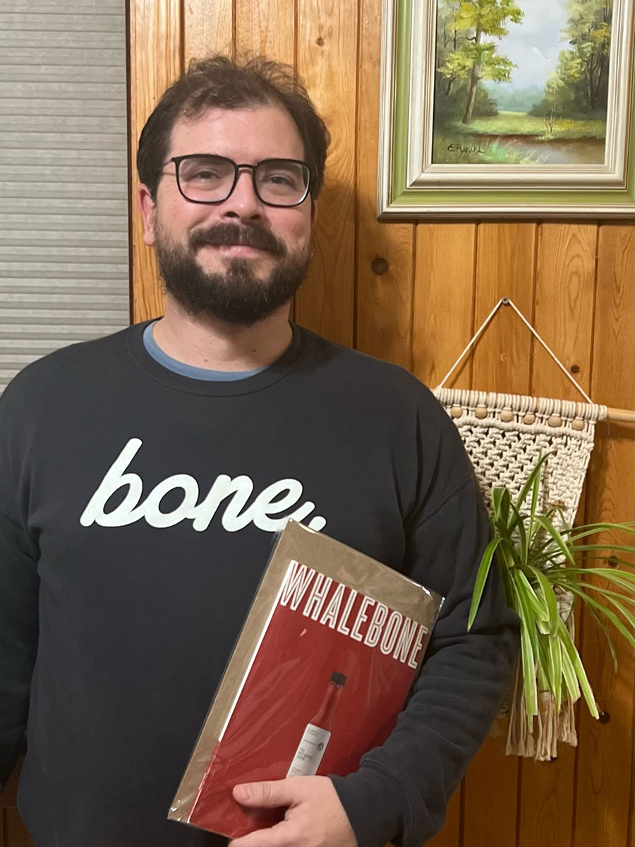Photo of Matthew Wisotsky, a brown-haired man with a thick beard and mustache who wear black square-framed glasses. Mathew is wearing a gray "bone." shirt and holding The Hot Sauce Issue of Whalebone Magazine against his side with his left hand while facing the camera and smiling slightly.