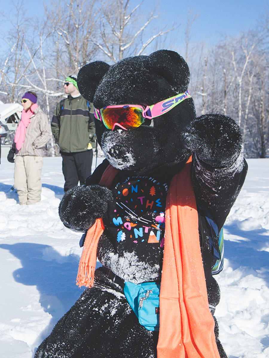 Photo of a person in a full-body black bear costume covered in snow at one of the WHP On The Mountain series events. The person in the bear suit is standing on the top of a white snow-covered ski hill and wearing bright pink and green snow goggles across the bear costume eyes and a light orange scarf while fake punching towards the camera. The person is also wearing black "On The Mountain" t-shirt with orange, blue and pink lettering forming a circle in the center.