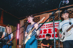 Feature photo taken front row from the audience of a band on stage mid-performance at one of The Wild Honey Pie PIzza Parties. A guyitar player in a white t-shirt is standing to the right, the lead singer is in blue jeans and a gray t-shirt and is playing guitar and singing into the mic in the middle, the drummer is in the back center and the keyboardist is on the left of the stage in blue overalls playing a black keyboard.