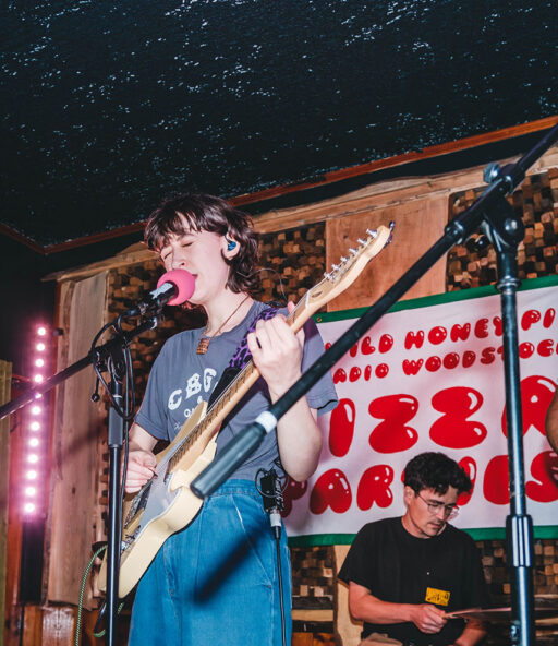 Feature photo taken front row from the audience of a band on stage mid-performance at one of The Wild Honey Pie PIzza Parties. A guyitar player in a white t-shirt is standing to the right, the lead singer is in blue jeans and a gray t-shirt and is playing guitar and singing into the mic in the middle, the drummer is in the back center and the keyboardist is on the left of the stage in blue overalls playing a black keyboard.