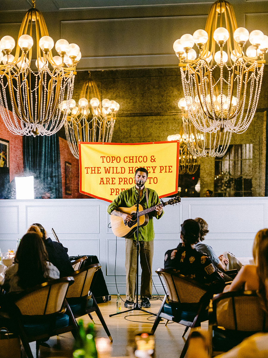 Photo of a man wearing a green button down and brown khakis singing on stage into a mic with a light brown acoustic guitar in his arms at a WHP Dinner Party. A large yellow banner lined with red and with red lettering that reads "TOPO CHICO & THE WILD HONEY PIE ARE PROUD TO PRESENT" is hanging behind him Large crystal chandeliers with yellow lights are hanging along the sealing and tables of people seated in brown and wicker wood chairs are sitting and watching the singer on stage.